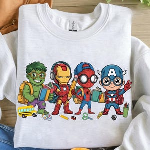 May include: A light gray sweatshirt featuring a colorful graphic of four cartoon superheroes, each with backpacks and school supplies. The characters include Hulk, Iron Man, Spider-Man, and Captain America, all ready for school.