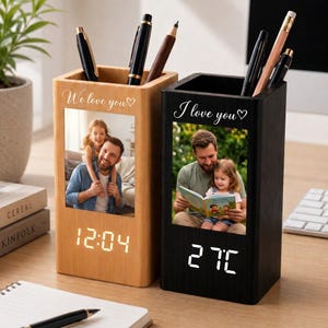 Peut inclure: Deux porte-stylos rectangulaires en bois avec affichage numérique. Le modèle en bois clair porte l'inscription "We love you" et une photo d'un homme et d'un enfant. Le modèle noir porte "I love you" et une photo d'un homme et d'un enfant.