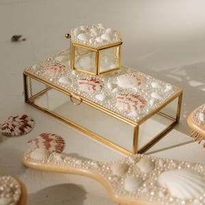 May include: A rectangular glass jewelry box with a gold metal frame, featuring a lid decorated with white pearls and seashells. A smaller hexagonal glass box with a gold frame sits on top. A hairbrush decorated with pearls and seashells is also visible.