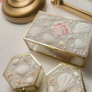 May include: Three decorative glass boxes with gold trim, adorned with seashells and faux pearls. The boxes are in various shapes, including rectangular and hexagonal, and are arranged on a white surface. The boxes are likely used for jewelry or trinket storage.