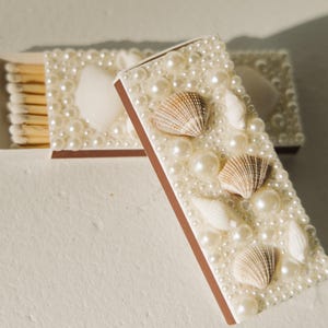 May include: Two decorative matchboxes, one open, adorned with faux pearls and seashells. The matchboxes have a brown border and contain wooden matches. The design creates a coastal or beach-themed aesthetic.