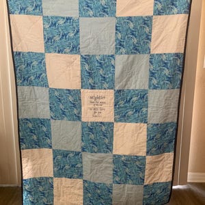 Può includere: Una trapunta patchwork blu e bianca con una citazione ricamata al centro: "Più potente delle onde del mare è il suo amore per te." Giovanni 4:18