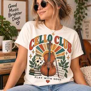 以下が含まれることがあります： 「CELLO CLUB」と「PLAY WITH SOUL」の文字がプリントされたクリーム色のTシャツ。デザインには、茶色のチェロ、弓、楽譜、積み重ねられた本、緑の葉、そしてオレンジとティールブルーのサンバースト模様が描かれています。