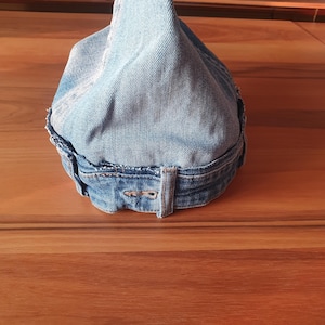 May include: A light blue denim hat with a pointed crown. The hat is made from recycled denim and features a belt loop detail at the back. The edges of the hat are frayed, giving it a vintage look.