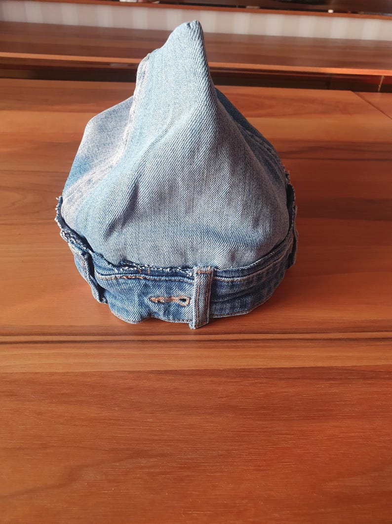 May include: A light blue denim hat with a pointed crown. The hat is made from recycled denim and features a belt loop detail at the back. The edges of the hat are frayed, giving it a vintage look.