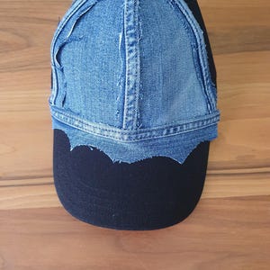 May include: A baseball cap featuring a black fabric base with a blue denim overlay on the front panels. The denim section has frayed, raw edges and a scalloped hemline that sits above the solid black brim of the hat.