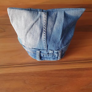 May include: A denim fabric container, possibly a bag or basket, crafted from repurposed blue jeans. The design incorporates the original waistband, belt loops, and seams of the jeans, showcasing a unique, upcycled aesthetic. The container sits on a wooden surface.