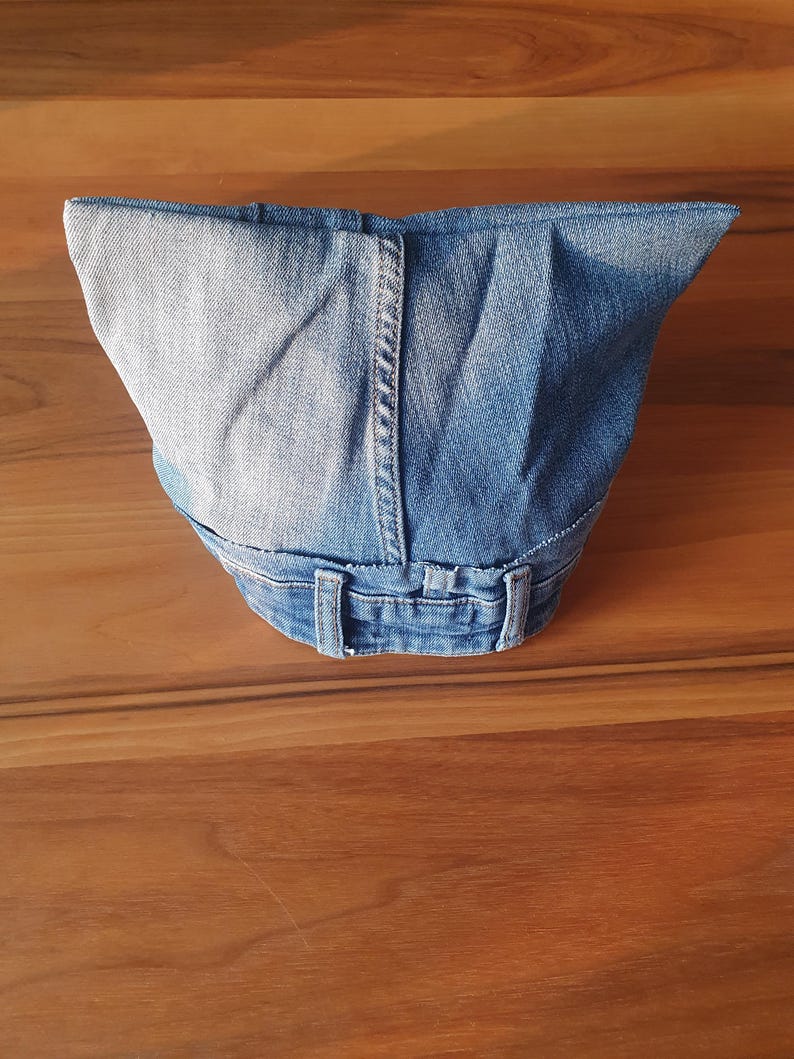 May include: A denim fabric container, possibly a bag or basket, crafted from repurposed blue jeans. The design incorporates the original waistband, belt loops, and seams of the jeans, showcasing a unique, upcycled aesthetic. The container sits on a wooden surface.