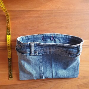 May include: A denim basket made from recycled jeans, showcasing the waistband, belt loops, and seams. A yellow measuring tape is positioned to the side. The basket is a light blue color and sits on a wooden surface.