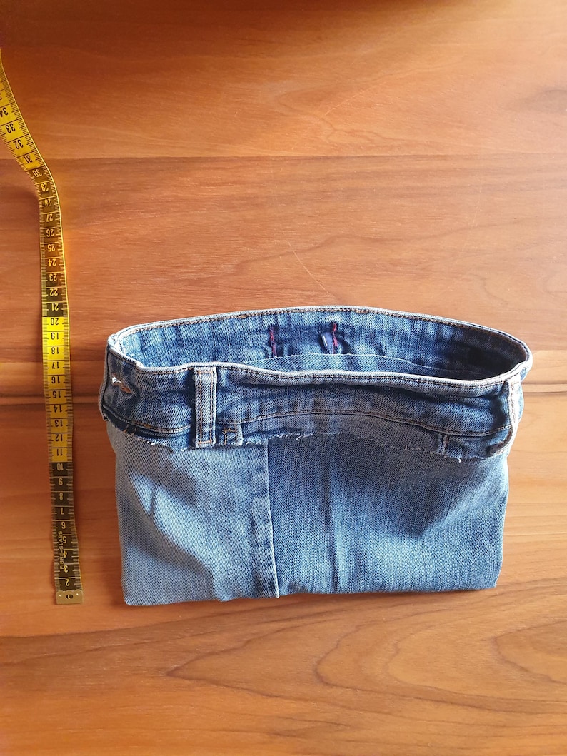 May include: A denim basket made from recycled jeans, showcasing the waistband, belt loops, and seams. A yellow measuring tape is positioned to the side. The basket is a light blue color and sits on a wooden surface.