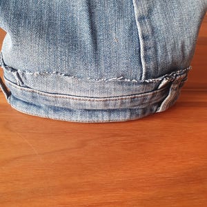 May include: Close-up of light blue denim fabric, showing the hem of a garment. The hem is folded over and stitched with white thread. The denim has a worn appearance.