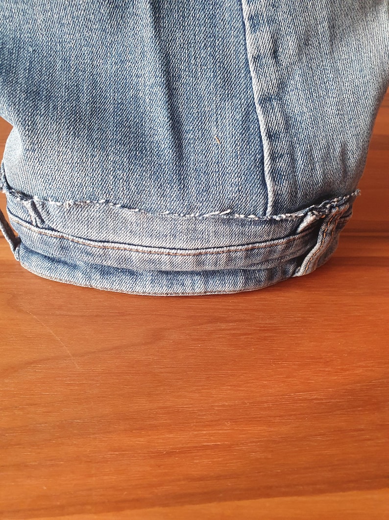 May include: Close-up of light blue denim fabric, showing the hem of a garment. The hem is folded over and stitched with white thread. The denim has a worn appearance.