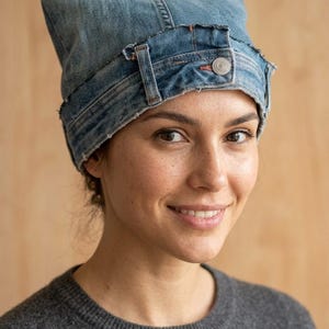 May include: A blue denim hat crafted from repurposed jeans, featuring a structured waistband with a visible metal button and belt loops. The design includes two pointed ears at the top, with frayed edges along the seams and a classic denim texture.