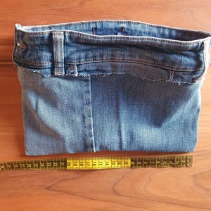May include: A handmade denim pouch crafted from repurposed jeans. The pouch features the original waistband, belt loop, and a raw-edge finish. A yellow measuring tape is placed next to the pouch, showing measurements in inches.
