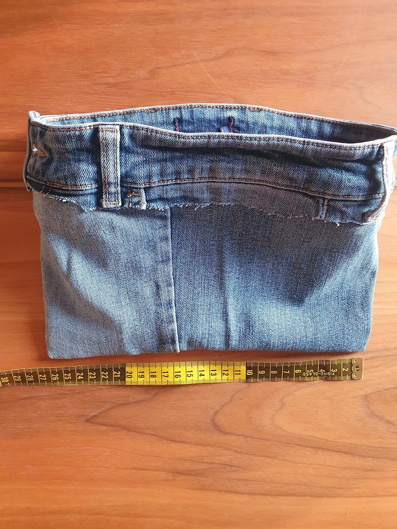 May include: A handmade denim pouch crafted from repurposed jeans. The pouch features the original waistband, belt loop, and a raw-edge finish. A yellow measuring tape is placed next to the pouch, showing measurements in inches.