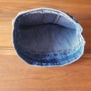 May include: A denim cap, viewed from the inside, showcasing the blue fabric and stitching details. The cap's brim is visible, and the interior reveals the construction and shape of the hat. The cap is set against a wooden surface.