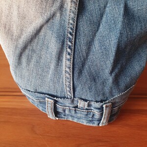 May include: Close-up of light blue denim jeans, showing the waistband, belt loops, and stitching details. The denim has a faded appearance, with lighter areas contrasting with the darker blue fabric. The jeans are folded.