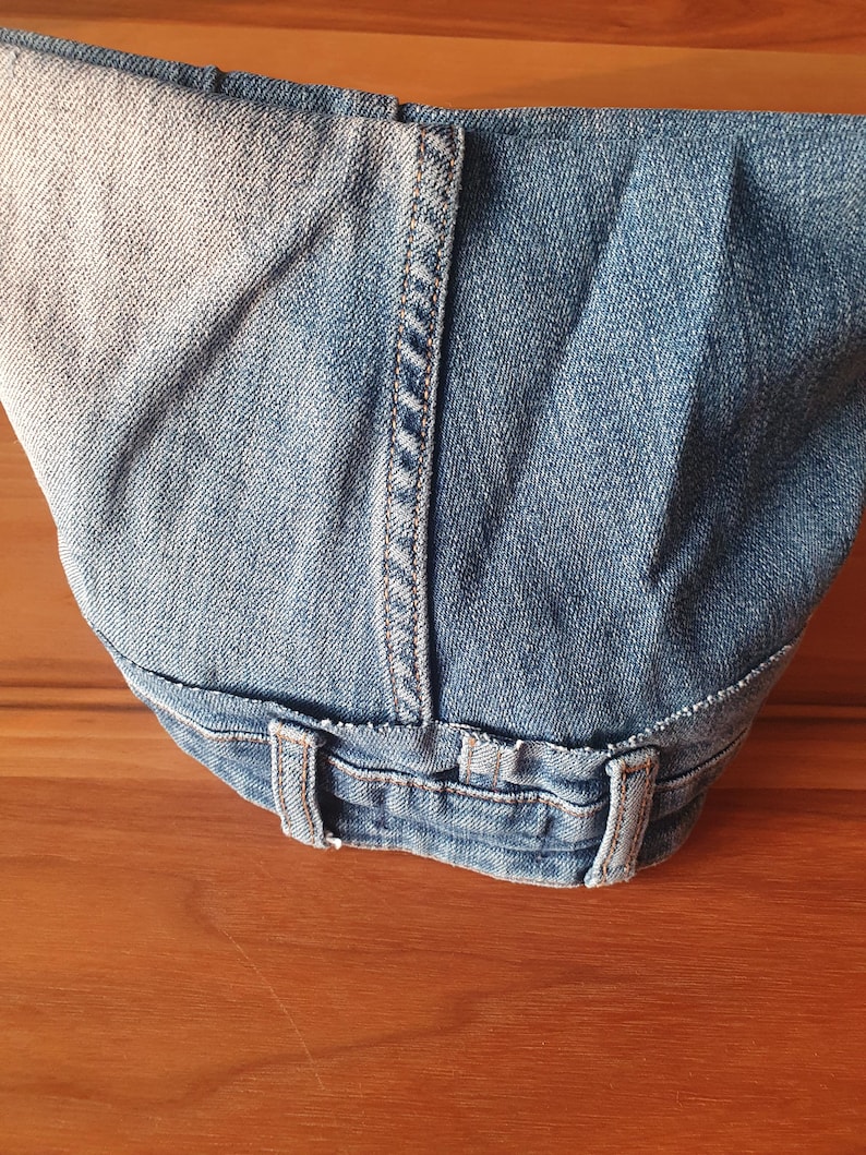 May include: Close-up of light blue denim jeans, showing the waistband, belt loops, and stitching details. The denim has a faded appearance, with lighter areas contrasting with the darker blue fabric. The jeans are folded.