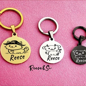 May include: Three metal keychains with engraved pet illustrations. The first is gold with a cat wearing a pirate hat, the second is silver with a cow, and the third is black with a dog. Each keychain has the name "Reese" engraved.