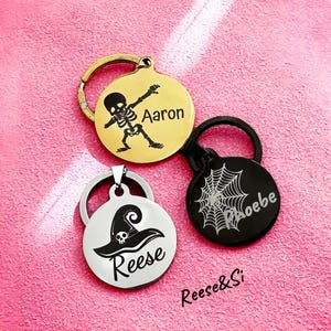 May include: Three round metal keychains with Halloween themes. The first keychain is gold with a skeleton in a dancing pose and the name "Aaron" engraved. The second keychain is black with a spiderweb design and the name "Phoebe" engraved. The third keychain is silver with a witch's hat and the name "Reese" engraved.