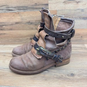 Puede incluir: Botines de cuero marrón con hebillas y correas. Las botas tienen un aspecto desgastado, con una mezcla de texturas y tonos marrones. El diseño incluye un puño doblado y un tacón bajo.