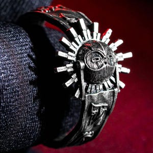Puede incluir: Un anillo plateado con un diseño detallado en forma de sol, con una cara central y puntas radiantes. El anillo tiene patrones intrincados y se encuentra sobre un fondo oscuro, resaltando su acabado metálico y artesanía.