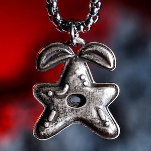 May include: A silver-colored pendant necklace featuring a stylized star design with a central hole and decorative details. The pendant is suspended from a matching chain, set against a blurred red and gray background.