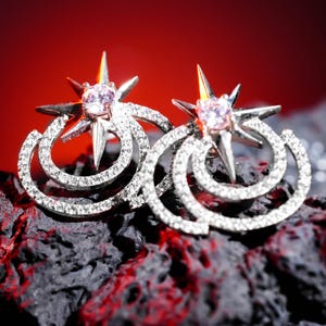May include: A pair of silver-toned stud earrings. Each earring features a starburst design with a central pink gemstone and is surrounded by a spiral of small, clear crystals. The earrings are set against a dark, textured background.