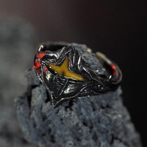 Puede incluir: Un anillo de plata con un dise&ntilde;o negro y amarillo. El anillo est&aacute; colocado sobre un fondo gris oscuro.