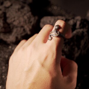 May include: A silver snake ring with a detailed design. The ring is worn on the index finger of a hand.
