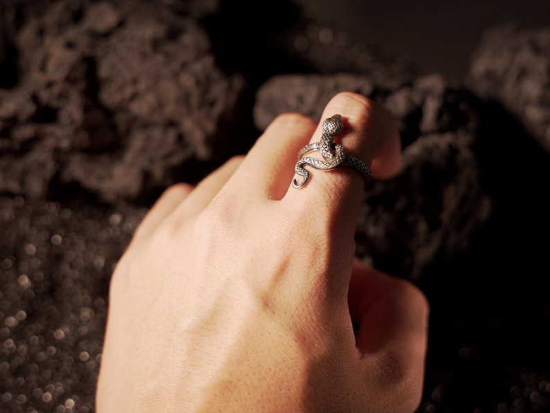 May include: A silver snake ring with a detailed design. The ring is worn on the index finger of a hand.