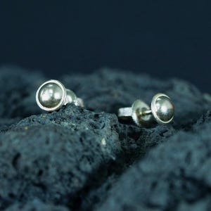 May include: A pair of silver stud earrings with a rounded, domed design. The earrings are set against a dark, textured background, highlighting their metallic sheen. The studs have a simple, minimalist style.
