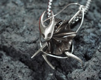 May include: A silver pendant necklace featuring a detailed, stylized depiction of the Knight from the video game Hollow Knight. The pendant is attached to a silver chain.