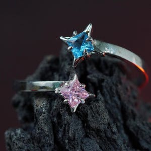 Könnte beinhalten: Zwei silberne Ringe mit blauen und rosa sternförmigen Edelsteinen. Die Ringe liegen auf einer dunkelgrauen, strukturierten Oberfläche.