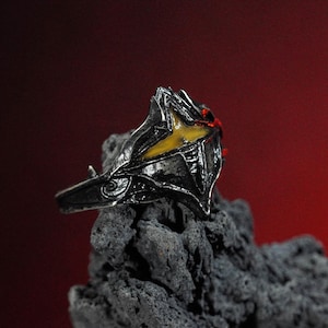 Puede incluir: Un anillo de plata con un dise&ntilde;o detallado que presenta una piedra preciosa amarilla y roja. El anillo est&aacute; colocado sobre un fondo gris oscuro.