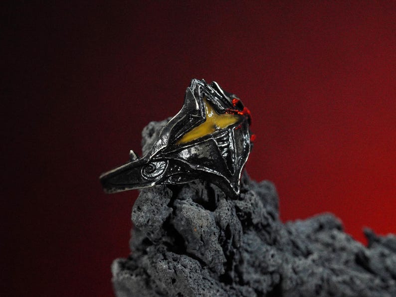 Puede incluir: Un anillo de plata con un dise&ntilde;o detallado que presenta una piedra preciosa amarilla y roja. El anillo est&aacute; colocado sobre un fondo gris oscuro.