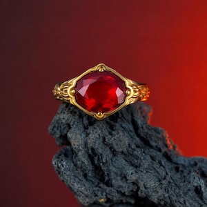 May include: A gold ring featuring a large, faceted red gemstone. The ring has an ornate, filigree design on the band and around the gemstone setting. It is displayed on a dark, textured surface against a red backdrop.
