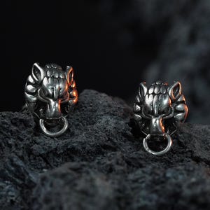 May include: A pair of silver wolf head stud earrings with a ring detail, presented on a dark, textured surface. The earrings feature detailed facial features and are set against a dark backdrop.
