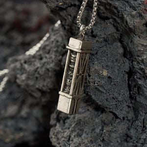 May include: A silver-colored pendant necklace featuring a rectangular, detailed charm. The charm has a cylindrical design with etched patterns and is suspended from a silver chain. The background is a dark, textured rock formation.