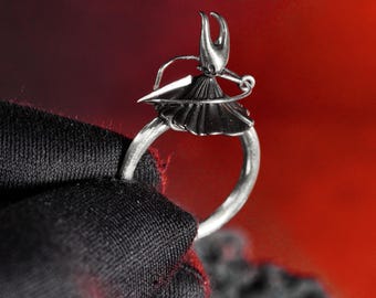 May include: A silver ring featuring a stylized figure with a headpiece, a skirt, and a sword. The ring is held against a dark fabric with a red background. The ring's design is intricate and detailed.