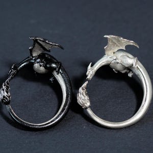 May include: Two dragon rings, one black and one silver, are displayed. Each ring features a dragon curled around the band, with the dragon's head facing a flame at the opposite end. The dragons have detailed wings and scales.