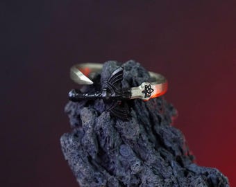 May include: A black and silver ring with a sword design. The sword is black and has a silver handle. The ring is set on a dark gray background.