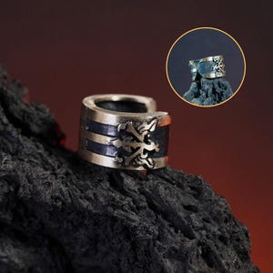 May include: A silver and black ear cuff with a detailed design. The cuff is made of metal and has a black enamel finish. The design features a stylised floral pattern.