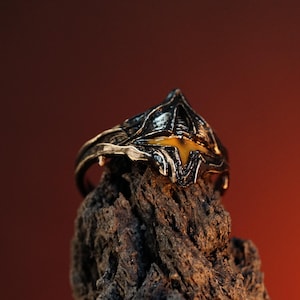 Puede incluir: Un anillo de plata con un dise&ntilde;o detallado de un casco con una piedra preciosa amarilla en el centro. El anillo est&aacute; sobre un trozo de madera &aacute;spera.