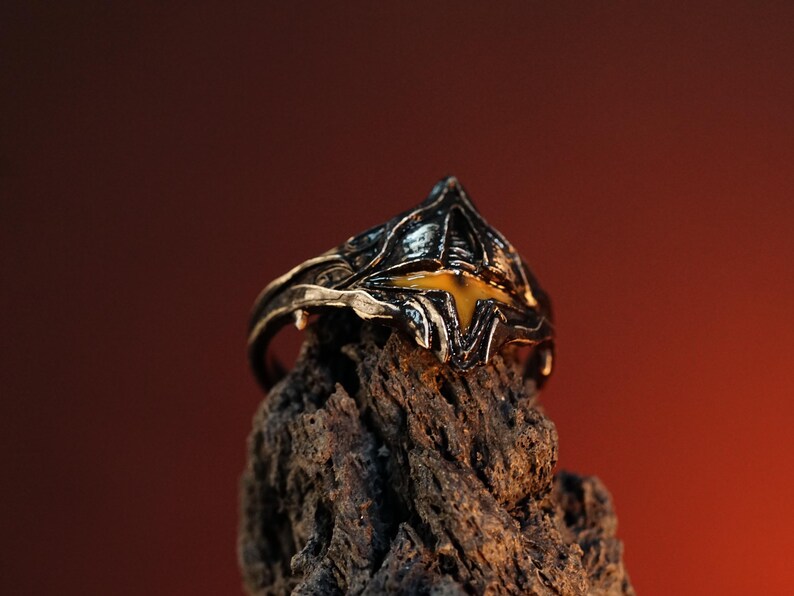 Puede incluir: Un anillo de plata con un dise&ntilde;o detallado de un casco con una piedra preciosa amarilla en el centro. El anillo est&aacute; sobre un trozo de madera &aacute;spera.