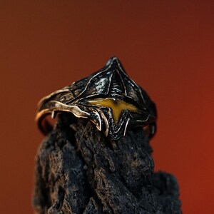 Puede incluir: Un anillo de plata con un dise&ntilde;o negro y amarillo. El anillo tiene forma de corona con una piedra amarilla en el centro.