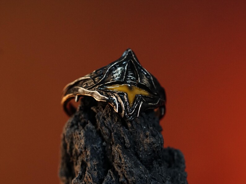 Puede incluir: Un anillo de plata con un dise&ntilde;o negro y amarillo. El anillo tiene forma de corona con una piedra amarilla en el centro.