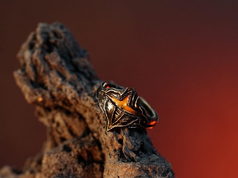 Puede incluir: Un anillo de plata con un dise&ntilde;o detallado que presenta una piedra amarilla en forma de estrella. El anillo est&aacute; apoyado en un trozo de madera desgastada.