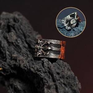 May include: A silver ring with a detailed design featuring a skull and crossbones. The ring is wide and has a textured finish.