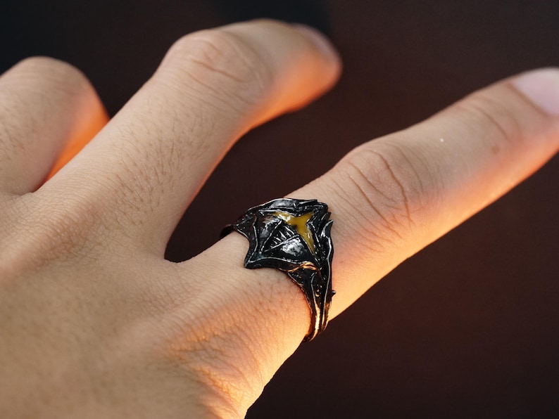 Puede incluir: Un anillo negro con una piedra amarilla en el centro. El anillo est&aacute; dise&ntilde;ado con una forma afilada y angular.